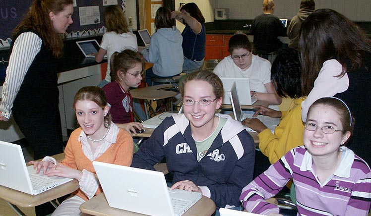 Three girls smiling behind laptops in a classroom setting.