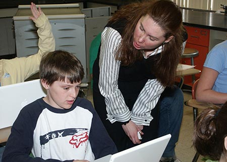 A teacher helps a student at a laptop in a classroom.